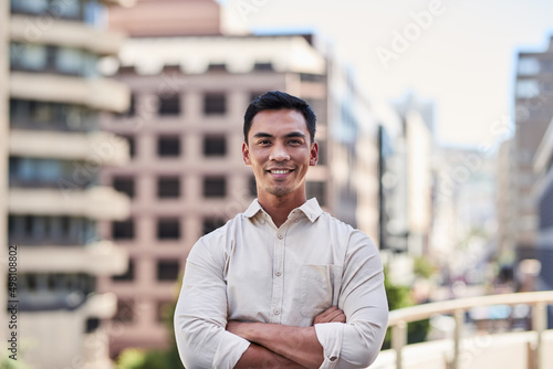 A young attractive Asian man stands in front of city buildings with arms crossed photo