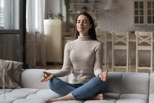 Peaceful calm millennial yogi girl meditating with closed eyes on home couch, keeping hand zen fingers, serene face, doing yoga, mental exercise, practicing mindfulness