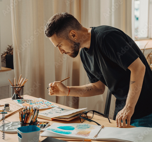 A creative person is standing over a desktop with a brush in his hand. A young artist makes a sketch with a brush in the studio. Creative studio, lifestyle, the process of creating a work of art photo
