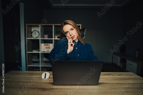 busy young woman sitting at home on laptop at work and talking on the phone, looking at the camera with a serious face