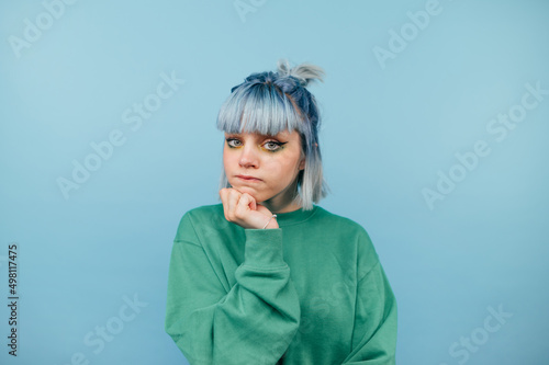 Pensive young lady with blue hair stands on a colored background and looks into the camera with a serious face. Isolated.