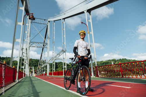 Handsome young male cyclist in sports outfit stands with bicycle on the street on the background of the bridge.
