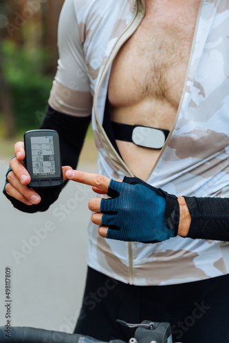 A male cyclist with a heart rate monitor on his chest stands with a bicycle outside the city during training and points his finger at the bicycle computer. © bodnarphoto