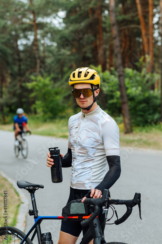 Handsome athletic man in outfit stands with a bicycle outside the city in the woods on the road with a bottle of water in his hand and looks at the camera.