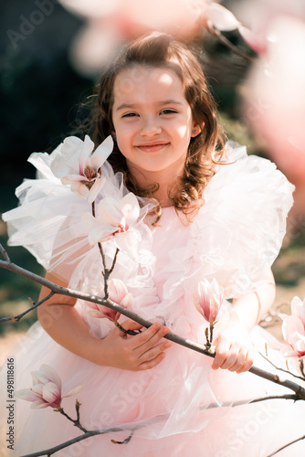 Beautiful smiling kid girl 4-5 year old wear stylish princess dress smell magnolia flower over blooming tree in garden outdoor. Spring season. Childhood. Springtime. Happy child with floral nature.
