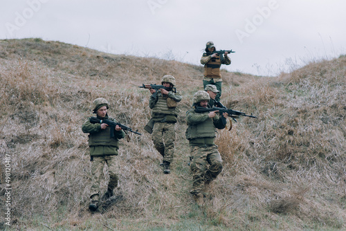 Team of five Ukrainian soldiers aims from kalashnikov assault rifles to russian enemy against background of hill. photo