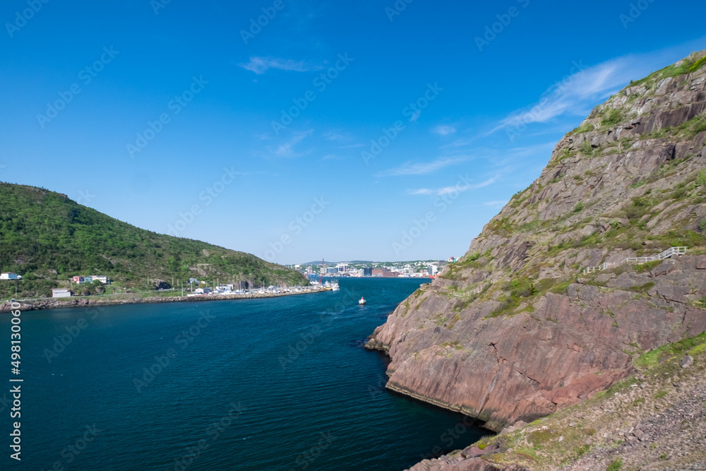 A footpath, Signal Hill hiking trail or path along a hillside. The cliff is rocky with grass patches. The city of St. John's, Newfoundland, is in the background on a sunny day. The sky is bright blue.