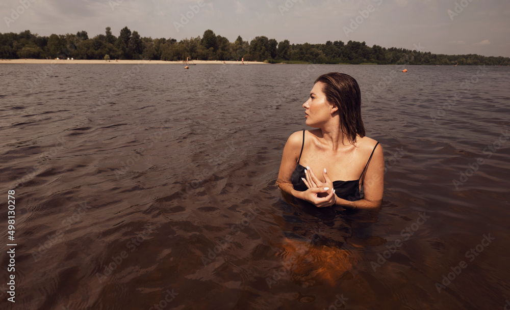 A young wet woman in a dark dress stands in the water. The woman swims in the river. brown tint A young wet woman in a dark dress stands in the water. The woman swims in the river. brown tint