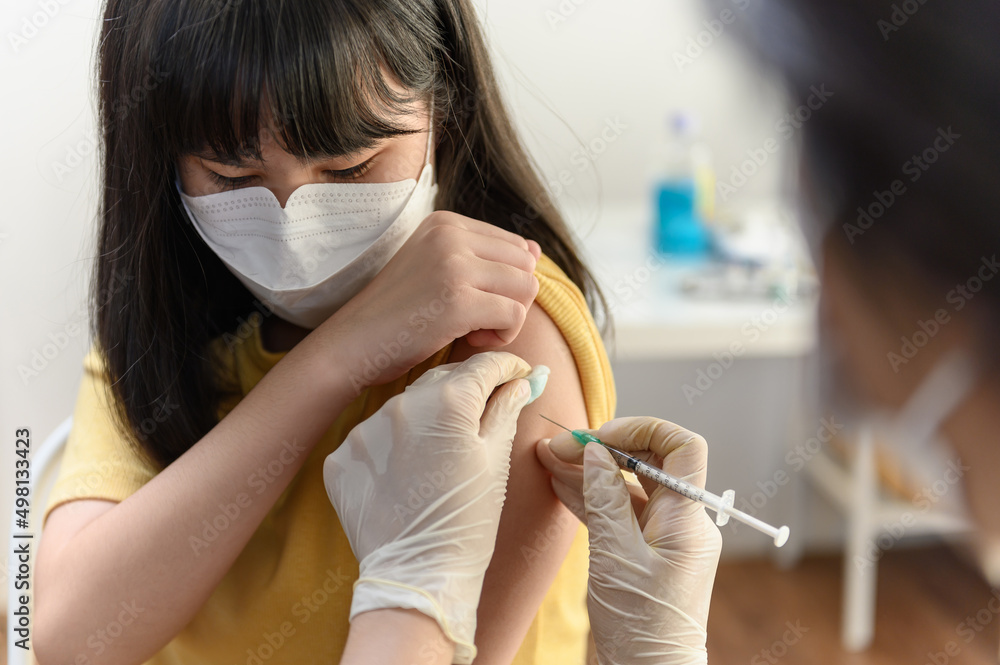 Asian woman nurse injecting covid-19 vaccine to young Thai girl patient wearing face mask in clinic or health care center. Coronavirus pandemic protection or kids health care medical concept.