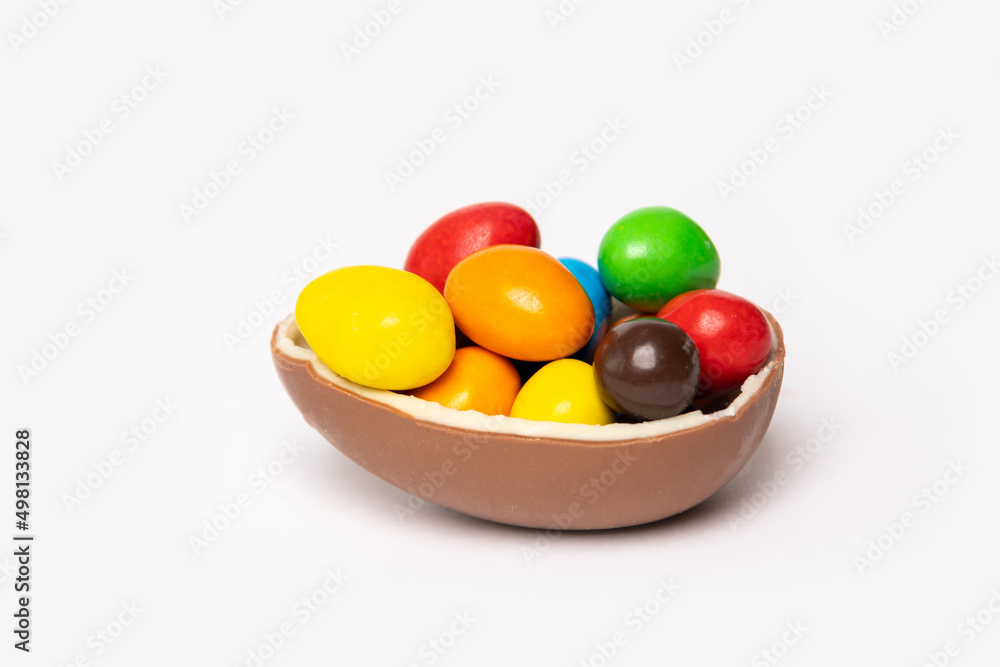 Cracked chocolate easter egg with colorful small round candies on white background, copy space, side view. Chocolate treat for kids. Easter concept. Cracked chocolate easter egg with colorful small round candies on white background, copy space, side view. Chocolate treat for kids. Easter concept.