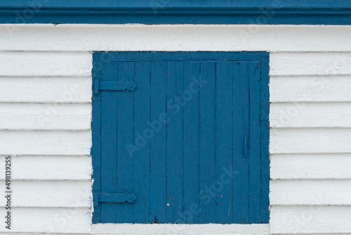 A vibrant royal blue wooden shutter door on a white wood wall. The exterior of the building is covered in white horizontal beveled clapboard siding. The boards on the window are vertical with hinges. 