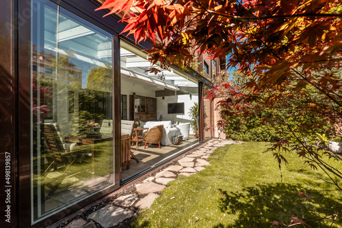 Closed terrace of brown aluminum and glass with large sliding doors, wooden and wicker furniture inside, a walk of stone slabs and a beautiful garden with lawn and ornamental trees