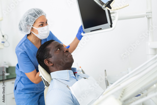 Confident asian female stomatologist showing teeth radiography result on computer screen to african american male patient, discussing medical treatment at clinic..