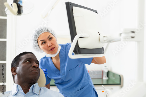 African-american man sitting on dental chair. Asian woman dentist pointing at display.
