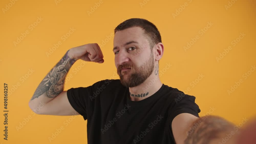 Studio shot of a young caucasian gangster-like tattooed man flexing his biceps and giving the eye over yellow background. High quality 4k footage Studio shot of a young caucasian gangster-like tattooed man flexing his biceps and giving the eye over yellow background. High quality 4k footage