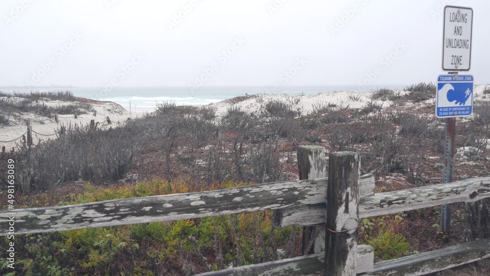 Ocean beach sandy dunes, Monterey, California misty coast, USA. Foggy rainy autumn, winter weather, grey cloudy sky. Trail path on shore, cold sea waves. Warning sign, tsunami hazard. Danger caution.