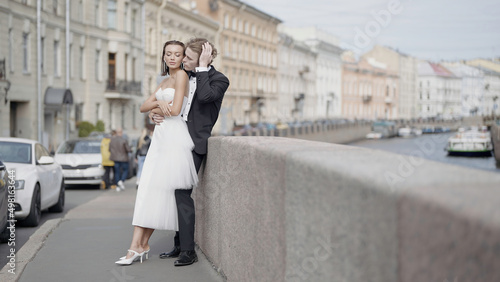 Beautiful newlyweds. Action. A couple of lovers, the bride in a tight white dress and the groom in a suit with long hair pose on the street next to the embankment and beautiful historical buildings in