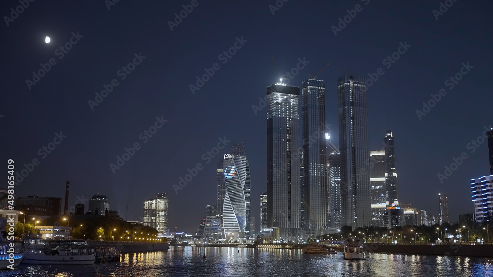 Late evening cityscape with Moscow city skyscrapers on dark blue night sky. Action. Beautiful river and glowing buildings with a shining moon.