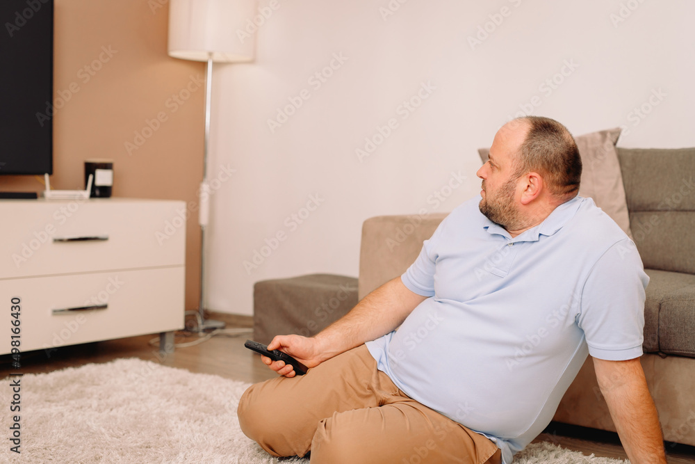Fototapeta premium Side view photo of a sibgle man sitting on the carpet at home near the sofa holding in his hands tv remote and watching tv.