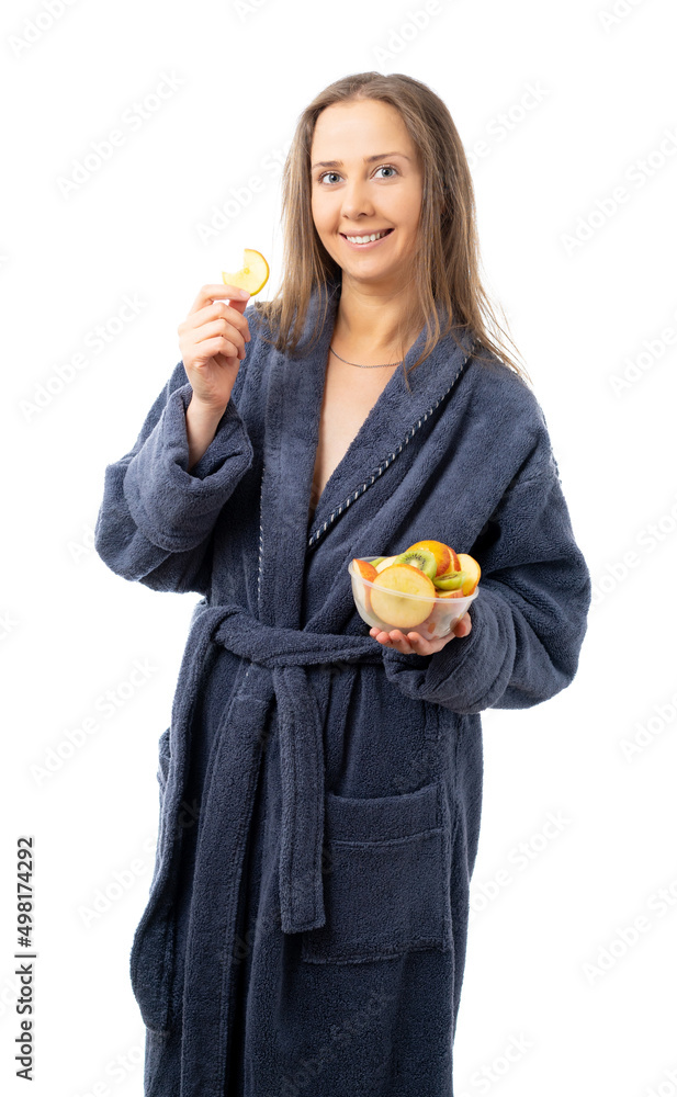 A woman in a gray terry bathrobe is a plate with sliced fruit and bites off an apple slice. isolated. A woman in a gray terry bathrobe is a plate with sliced fruit and bites off an apple slice. isolated.