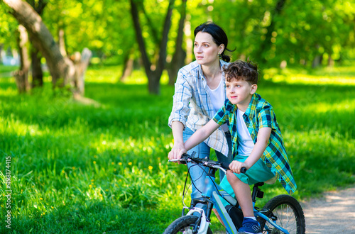 Mom helping her son learn to ride a bike in a summer park.