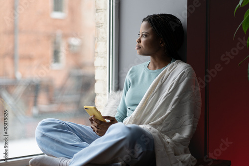 Cuadro en lienzo Focused African American woman covered with blanket sitting on windowsill, holding a phone, thinking about something