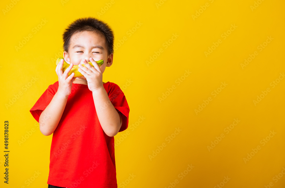 Happy portrait Asian child or kid cute little boy attractive smile wearing red t-shirt playing holds banana fruit, studio shot isolated on yellow background Happy portrait Asian child or kid cute little boy attractive smile wearing red t-shirt playing holds banana fruit, studio shot isolated on yellow background