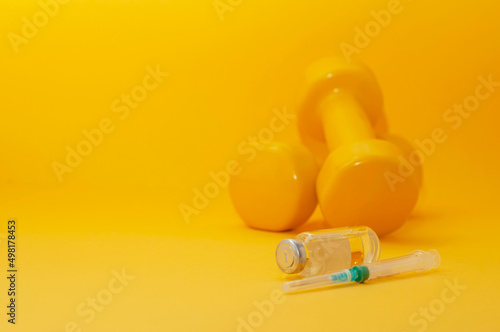 syringe and a jar of liquid lie in front of the dumbbells on a yellow background, copy space. the concept of doping in sports, steroids, testosterone and other drugs banned in sports.