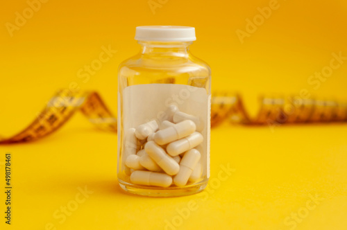 glass jar with tablets in capsules stands in front of a measuring tape on a yellow background, a horizontal snapshot. The concept of weight loss, fat burner, vitamins, sports nutrition.