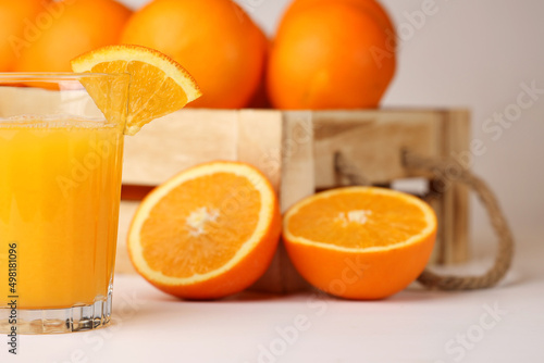 A glass of fresh orange juice, a slice of an orange on it, wooden box with rope handles full of oranges, one sliced orange on the side