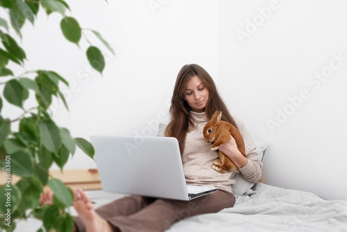 Caucasian brunette woman,girl with long hair sitting on bed in front of laptop with cute little rabbit,pet in white scandinavian interior.Distance education,work from home,watching film.Copy space