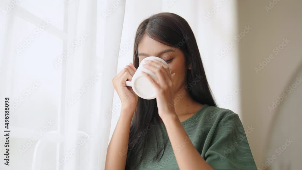 Young asian woman standing beside window and holding mug in bedroom at home, She drinking milk after wake up in the morning Young asian woman standing beside window and holding mug in bedroom at home, She drinking milk after wake up in the morning