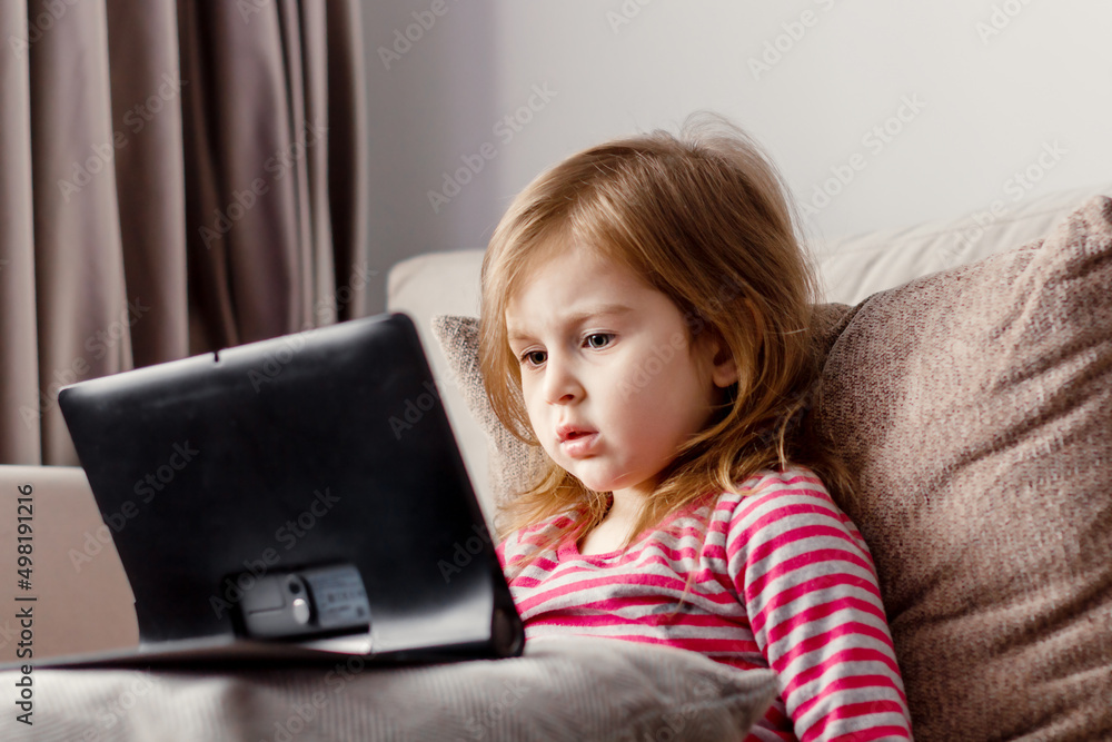 Cute toddler girl lying on beige couch with decorative pillows, holding tablet pad laptop gadget. Leisure moments at home with modern interior in neutral colors. Cute toddler girl lying on beige couch with decorative pillows, holding tablet pad laptop gadget. Leisure moments at home with modern interior in neutral colors.