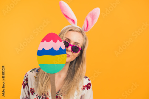 Happy Easter - a blonde young woman with Easter bunny ears holding a big wooden Easter egg holiday concept orange background medium closeup studio shot. High quality photo