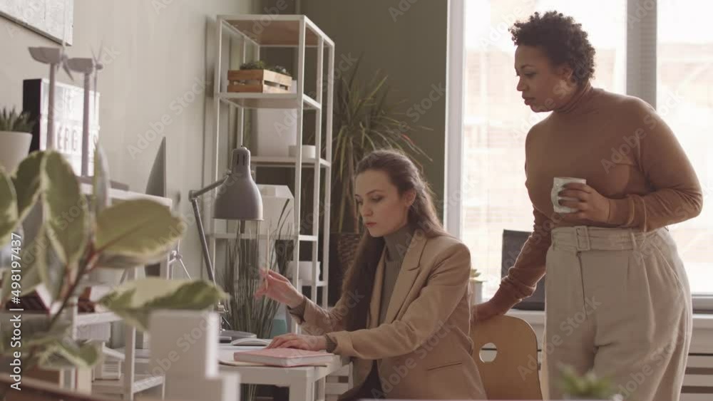 Medium slowmo of two young diverse female engineers working on project of renewable energy drawing paper plan and having discussion in office Medium slowmo of two young diverse female engineers working on project of renewable energy drawing paper plan and having discussion in office