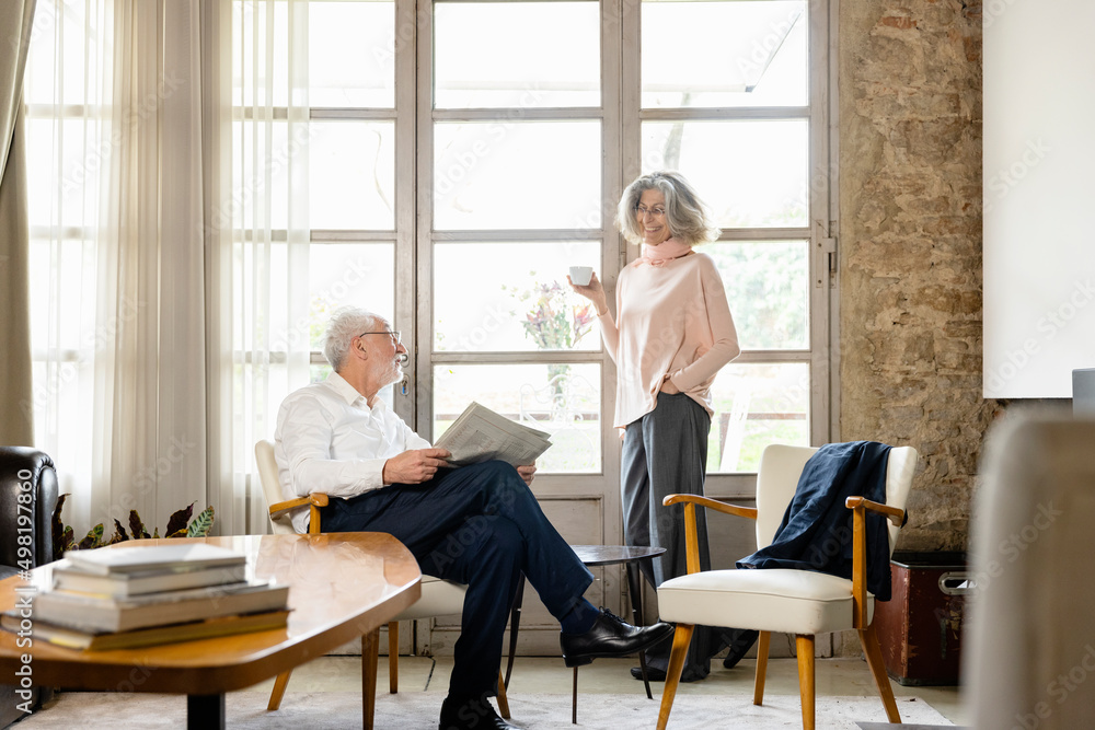 Smiling senior woman holding coffee cup talking with man in living room at home Smiling senior woman holding coffee cup talking with man in living room at home