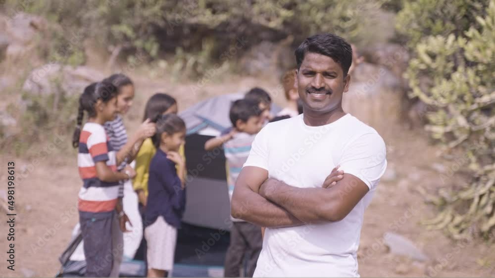 smiling Teacher standing with crossed arms in front camping tent with kids by looking at camera during summer camp - concept of mentorship, confident and supportive. smiling Teacher standing with crossed arms in front camping tent with kids by looking at camera during summer camp - concept of mentorship, confident and supportive.