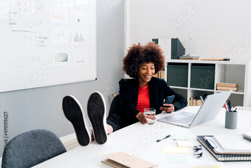 Happy businesswoman sitting with feet up on desk and using mobile phone in office photo