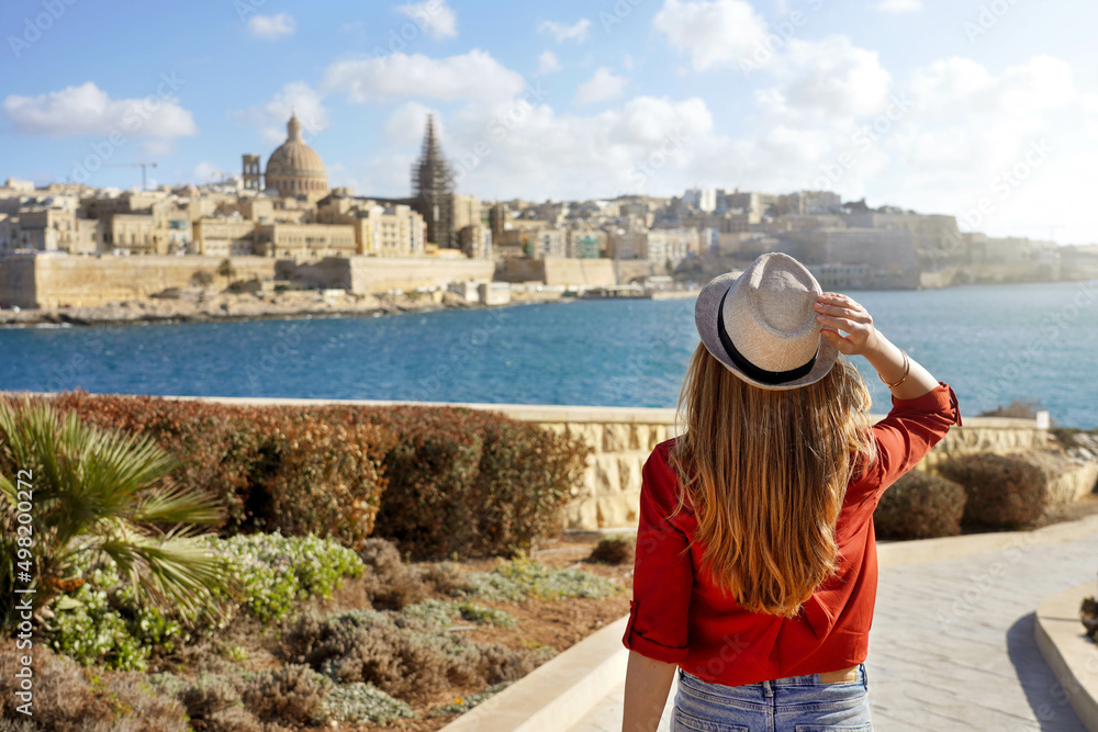 Beautiful young woman with hat goes down along Malta waterfront looking at stunning panoramic view of Valletta, Malta