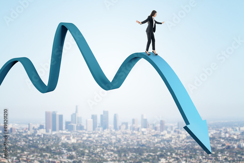 Attractive european businesswoman balancing on abstract falling blue arrow on sky and city background. Crisis, inflation and finance concept.