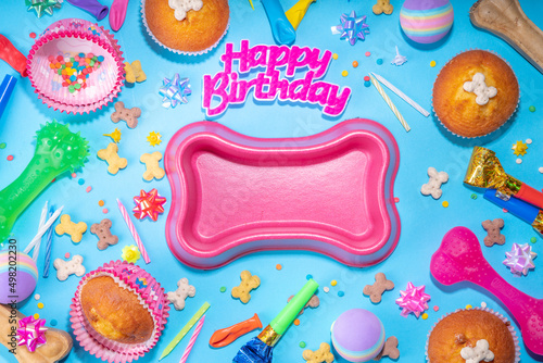 Pet happy birthday background with set different dogs snack, food and toys, bone-shaped dog bowl, birthday cupcakes with happy birthday candles and accessories. Flatlay on blue background top view