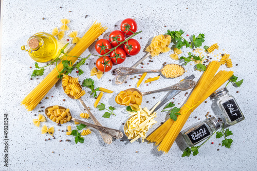 Various dried spaghetti, noodles, pasta set in spoons on white table background. Different shapes types raw italian pasta with herbs, olive oil, tomatoes, spices for cooking top view copy space