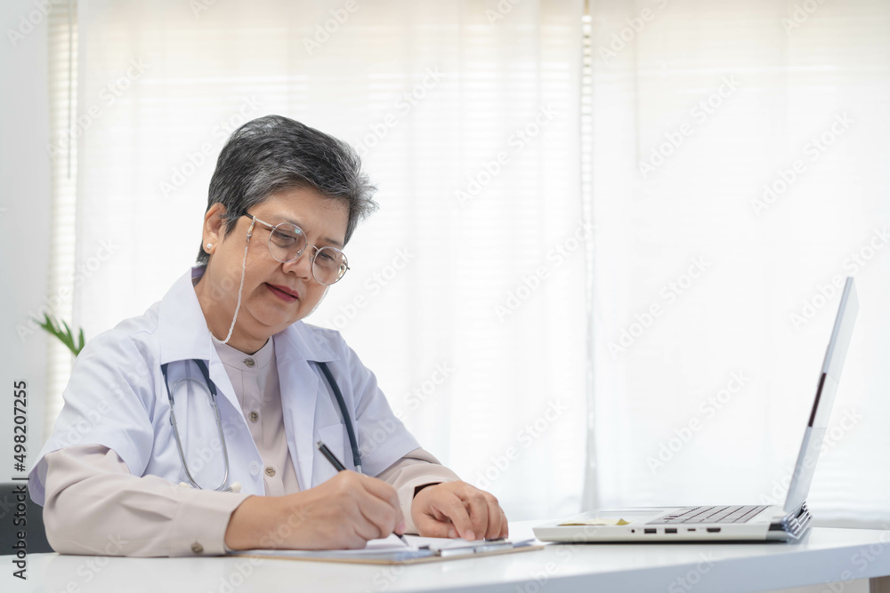 Senior asian physician, general woman doctor wearing glasses and white uniform, mature female with stethoscope, working on laptop computer, sitting in medical office. Healthcare and medical.