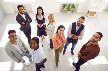 Diverse group of happy young people looking up and smiling at camera. Team of satisfied multiracial company employees standing in modern office all together. High angle indoor shot, view from above
