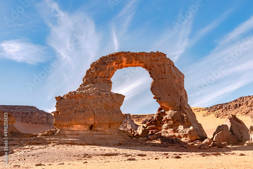 Amazing view in the Sahara desert of Tadrart in Djanet city, Algeria. With colorful orange sand, rocky mountains and an unbelievable arc shaped stone. Blue cloudy sky in background. 