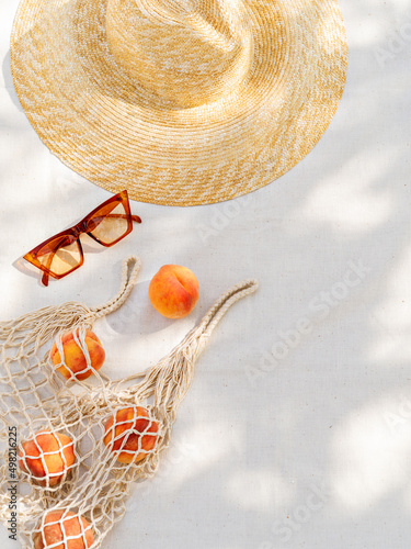 Vintage inspired background with straw hat, female sunglasses and shopper bag with peaches on white towel. Minimalist summer vacation creative still life ​for fashion blog, web, social media, stories.