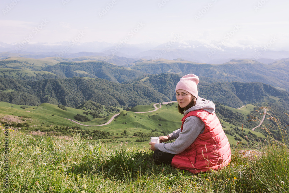 young woman is sitting against background of mountains and drinking tea from cup. travel, hiking, journey, outdoor recreation young woman is sitting against background of mountains and drinking tea from cup. travel, hiking, journey, outdoor recreation