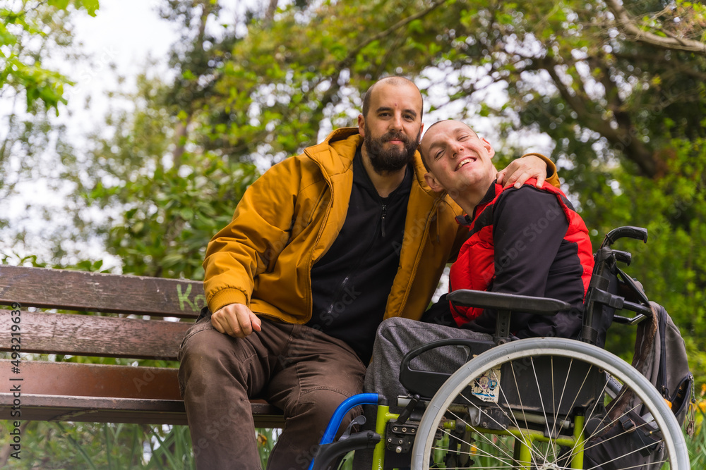 Portrait of a paralyzed young man in a wheelchair with a friend on a bench in a public park in the city Portrait of a paralyzed young man in a wheelchair with a friend on a bench in a public park in the city