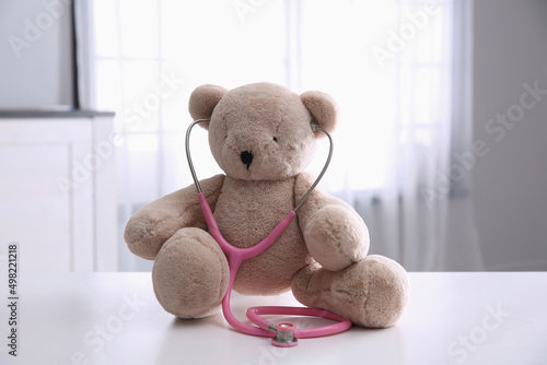 Cute teddy bear with stethoscope on white table indoors. Children's hospital