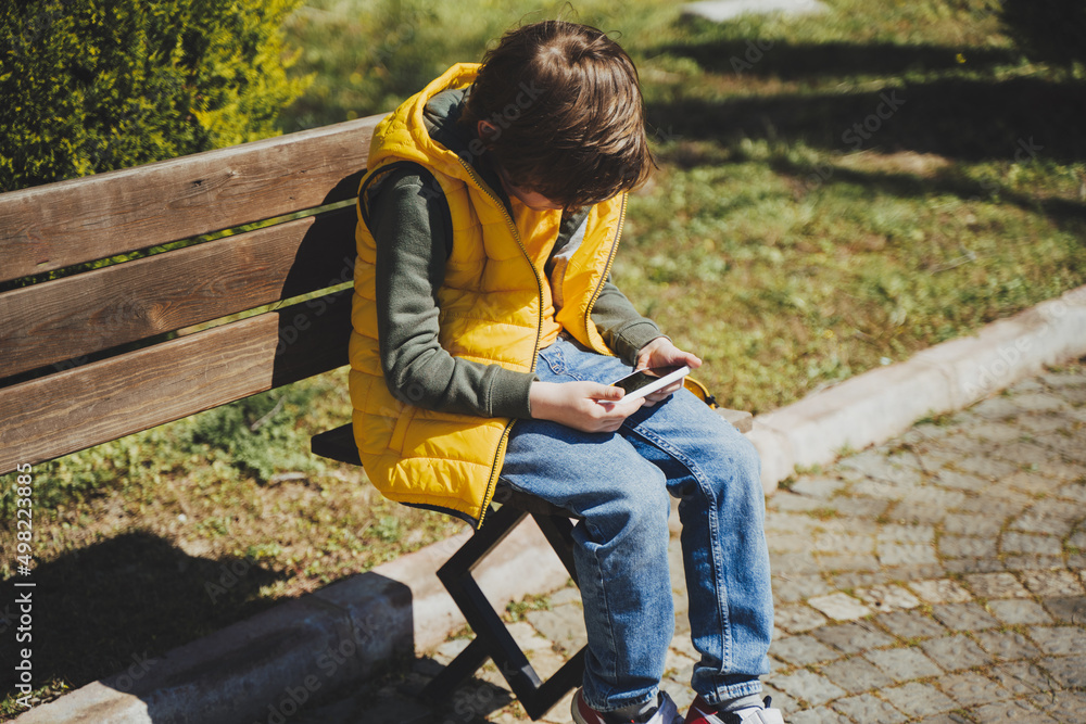 Schoolboy plays game his cell phone sitting on bench in the city park. Boy kid in yellow vest and green hoodie uses mobile gadget to chat with friends while outdoors in the garden during sunny day. Schoolboy plays game his cell phone sitting on bench in the city park. Boy kid in yellow vest and green hoodie uses mobile gadget to chat with friends while outdoors in the garden during sunny day.
